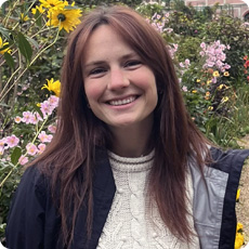 Andrea Mahoney wearing a white cable-knit sweater layered under a dark jacket is standing outdoors in a garden filled with colorful flowers. Bright yellow sunflowers and pink blossoms are visible in the background, along with green foliage. The setting appears vibrant and natural.