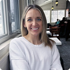 Kelsey MacNeil with shoulder-length blonde hair wearing a white ribbed sweater sits indoors near a large window in a cozy café or restaurant. The background features wooden chairs, hanging lantern-style lights, and other patrons seated at tables. Outside the window, a snowy winter scene is visible, adding to the warm, relaxed atmosphere.