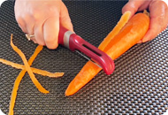A close up view of a person's hands peeling a carrot with a red vegetable peeler. There is a small pile of peelings on the surface.