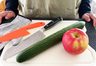 A cutting board with an apple, cucumber, knives and a safety glove laying on a table in front of a person.