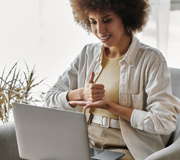 An African American woman signs with her hands while sitting in a chair and using a laptop.