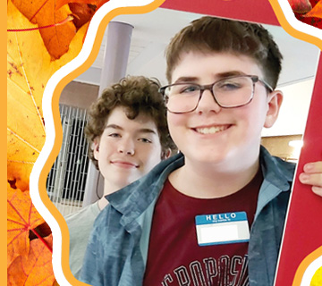 Two teenagers pose and smile for a photo, one wearing glasses and a name tag, with an autumn leaf-themed border around the image.