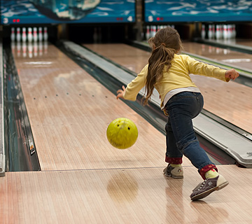 A child in a yellow shirt bowls a yellow ball down a lane at a bowling alley.