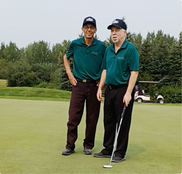 Gerry and Chris stand on a putting green with a golf cart in the background. Gerry holds a putter in his left hand.