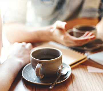 Two people have a discussion at a wooden table with notebooks and coffee cups, one person gesturing with a hand.