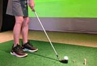 A person holding a golf club prepares to hit a ball on an indoor green with a virtual golf course projected on a screen.