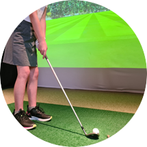 A person holding a golf club prepares to hit a ball on an indoor green with a virtual golf course projected on a screen.