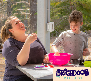 An adult and child are sitting at a table outdoors, engaged in an activity with a pink bowl. The logo on the image reads Brigadoon Village.