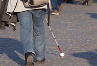 A man in black shoes, blue jeans and a light beige jacket with a light coloured backpack walks with a white cane down a sidewalk.