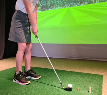 A person holding a golf club prepares to hit a ball on an indoor green with a virtual golf course projected on a screen.