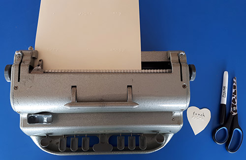 Perkins Brailler, braille paper, marker, scissor, small paper heart with braille/print message.