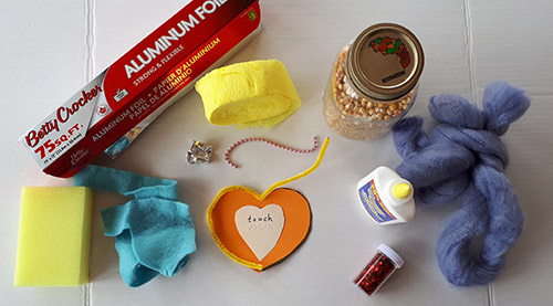 Posterboard heart with braille/print message surrounded with pipe cleaner, thick fluffy yarn, felt, sponge, crepe paper, glitter, popcorn, string of beads, aluminum foil, and glue.