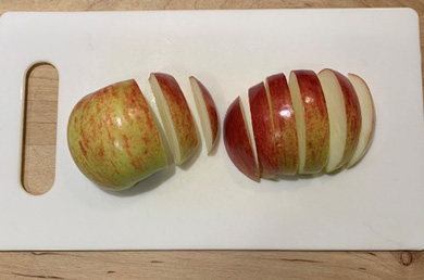 An apple cut in half with the peel facing up on a white cutting board. The apple on the left has been cut into two slices and the apple on the right has been cut into 6 slices.
