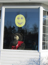 A house window taken from the outside with a yellow happy face cut-out and a teddy bear visible inside the window.