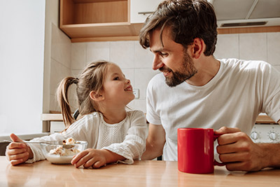 Dad and his little daughter are talking and smiling while having a breakfast in the kitchen at home.