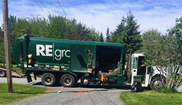 A green garbage truck on a residential street. A green compost bin and black garbage bag are on a lawn.