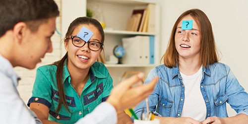 Three youths playing HedBanz with post-it notes on their foreheads. There is a boy talking and two girls with question marks on the post-it notes on their foreheads.