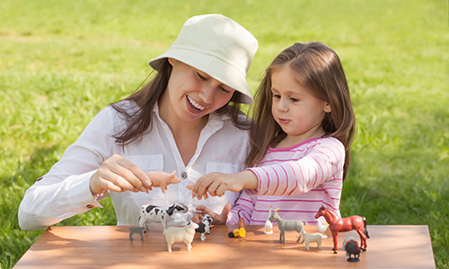 Mother and daughter playing with toy animals outside at a table on a sunny day