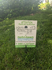 Pesticide notice sign, posted on a green lawn. Sign reads ATTENTION Pesticide Application and has information regarding application time and lawn care company's contact information.