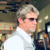 Patrick Friman sitting at a table in a resturant wearing a white shirt, grey vest and sunglasses