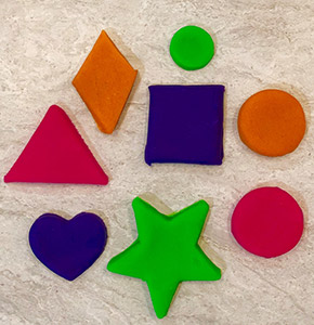 An array of playdough shapes including a large pink triangle, orange diamond, small green circle, large orange circle, large pink circle, large green star, purple heart, and large purple square