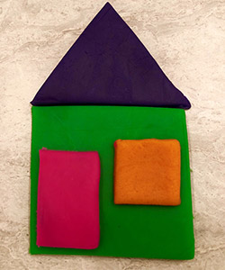 A house made of playdough shapes. The main base of the house is a very large green square. A large pink rectangle is placed on the left side of the green square to represent the door. A medium sized orange square is placed on the left side of the green square to represent a window.  A large purple triangle is placed above the base to represent the roof