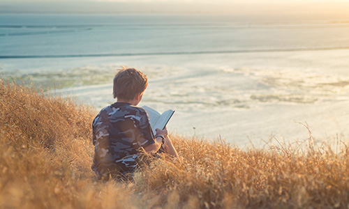 Young boy sitting on a hillside over looking the ocean while reading a book.