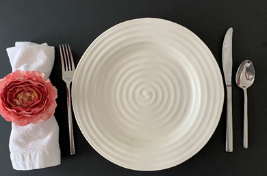 A white plate, silver utensils and a white napkin sitting on a black place mat. The napkin ring has a rose flower on top of it.