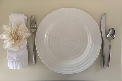 A table place setting with a white plate, white napkin and silver utensils on a white place mat. The napkin ring has a white flower on top of it.
