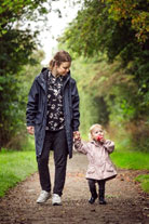 A mother and child walk hand in hand along a dirt path with trees on each side. The child is eating an apple.