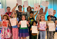 A group of diverse, smiling children proudly hold up their colorful activity sheets, showing off their completed work. They are standing in a bright room with a blue curtain in the background. The children are dressed in a mix of casual clothing, with some wearing vibrant colors and floral patterns. The atmosphere is lively and celebratory, indicating the completion of a fun group activity.