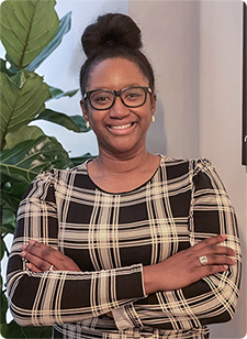 Melina Kennedy wearing glasses and a plaid dress smiles confidently with arms crossed. She stands in front of a leafy plant, creating a welcoming and professional tone.