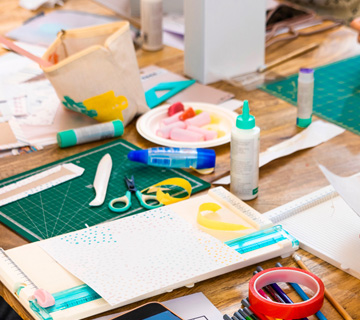 Assorted craft supplies on a wooden table, including scissors, glue, cutting mats, paper, tape, and small tools used for bookmaking or paper crafting.