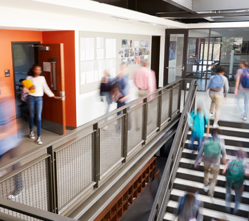 A busy school hallway with students in motion. Some walk along a staircase and others through a corridor, creating a dynamic and bustling atmosphere.
