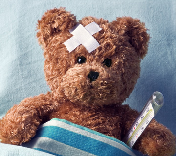 Teddy bear with a bandage on its head lies in bed, covered with a blue-striped blanket, holding a thermometer. The mood is caring and nurturing.
