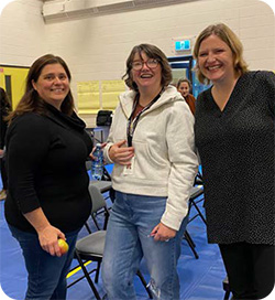 Three adults stand smiling together in a large indoor room set up for a professional learning session. They stand among rows of folding chairs, with other people talking in the background and flip-chart notes posted on the walls.
