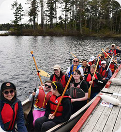 Campers at Brigadoon wearing red life jackets sit together in a long canoe. Many hold paddles upright and smile. The canoe floats on a calm lake.