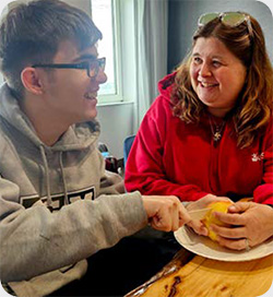 A young person and an adult sit at a table smiling and talking while working on a daily living skill. The young person uses a knife to cut a piece of fruit on a paper plate, while the adult provides guidance by holding the fruit steady.