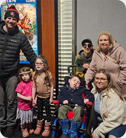 A group of adults and children pose together indoors near a movie theatre poster. One child sits in a wheelchair at the centre of the group, while other children stand close by holding hands.