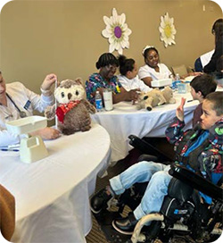 A healthcare professional sits at a table demonstrating a check-up on a stuffed owl during a teddy bear clinic activity. Several adults and children sit alongside the table, some holding stuffed animals, while a child using a power wheelchair raises a hand and watches attentively. The room is decorated with large paper flowers on the wall, creating a warm, welcoming setting.