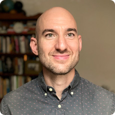 A bald man with a trimmed beard wearing a dark patterned button-up shirt stands in front of a bookshelf, smiling slightly at the camera.